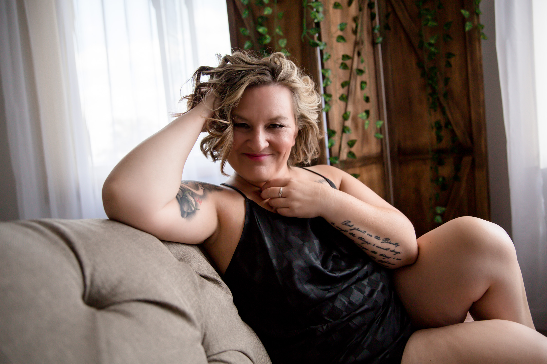A woman with short curly hair sitting on a couch with a cute flirty smile wearing a black pajama set for a photo session in Glencoe, Minnesota.